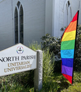 Photo of North Parish sign outside in front of church with a rainbow banner beside it.